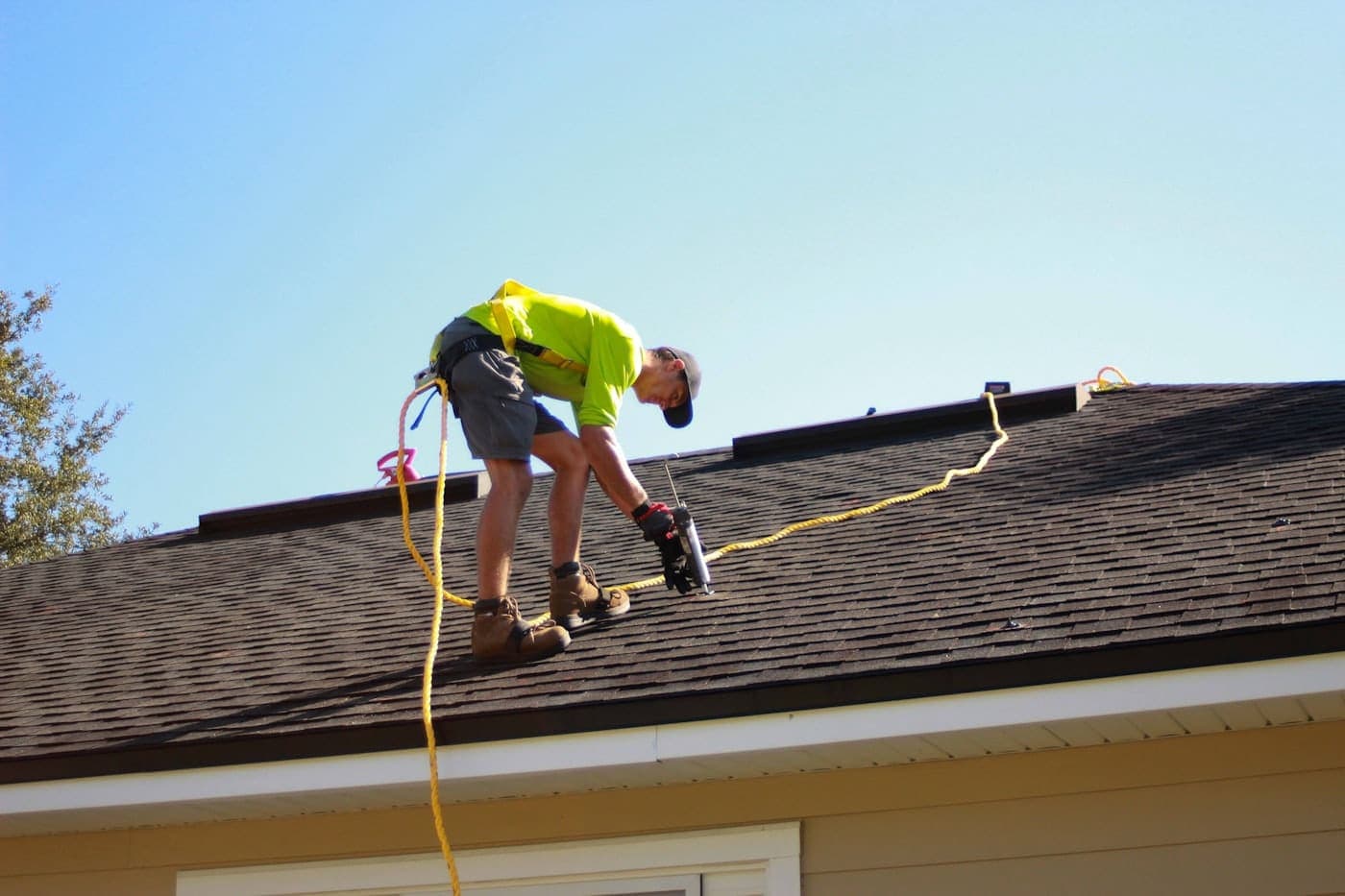 Roofer in a safety harness kneeling on a dark asphalt shingle roof, using a pneumatic nail gun to install shingles